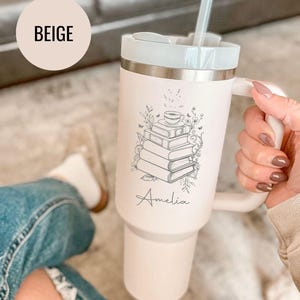 May include: Beige insulated tumbler with a white handle and lid. The tumbler features a black line drawing of a stack of books with a teacup on top, and the name "Amelia" in script. A clear straw is inserted in the lid.