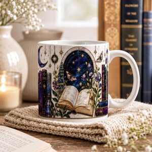 A white mug with a colorful, intricate design featuring a book, flowers, and a starry night sky, placed on a wooden surface next to a book and some flowers.