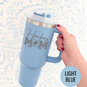 Engraved Hockey Mom tumbler with hockey stick and heart design; stainless steel insulated travel cup with handle; personalized drinkware for sports moms; custom cup for hockey games, practices, or gifts; durable large-capacity tumbler perfect for birthdays, holidays, and hockey season celebrations; stylish engraved design for everyday use.