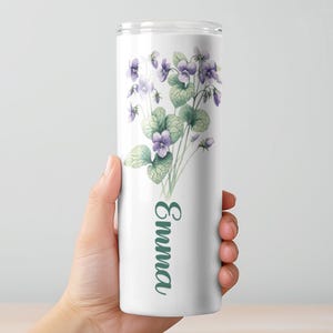May include: A white tumbler featuring a watercolor design of purple violets and green leaves. The name "Emma" is printed vertically in a green script font. The tumbler is held by a person.