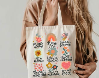 May include: A white canvas tote bag with colorful floral designs and inspirational messages. The text on the bag reads: "be kind always", "choose joy", "keep on blooming", "you matter", "focus on the good", "growth is a process", "the world needs you", "life is beautiful", and "you are loved".