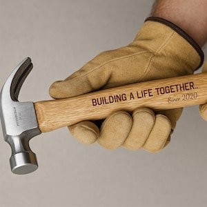 May include: A hammer with a wooden handle and a metal head is held by a gloved hand. The hammer is engraved with the words "BUILDING A LIFE TOGETHER.. Since 2020". The hammer head is marked "PITTSBURGH 16 oz".
