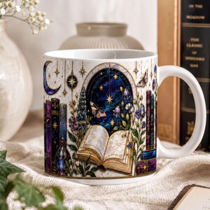 A white ceramic mug with a detailed illustration of a book, flowers, and a starry night sky on its surface, placed on a table with other objects such as books and a vase.