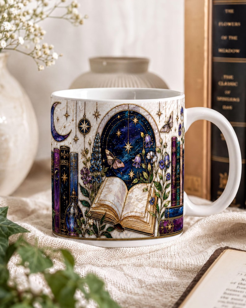 A white ceramic mug with a detailed illustration of a book, flowers, and a starry night sky on its surface, placed on a table with other objects such as books and a vase.