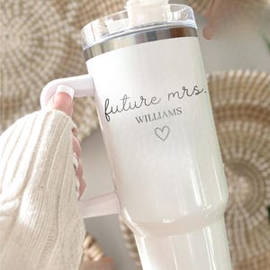 May include: White insulated tumbler with a handle, featuring the text "future mrs. Williams" in a script font, along with a heart symbol. The tumbler has a clear lid and a straw. The background is blurred.