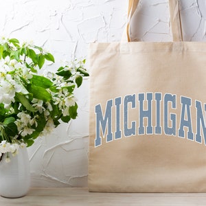 May include: A beige tote bag with the word "MICHIGAN" printed in blue on the front. The bag is sitting on a light wood surface in front of a white wall with a bouquet of white flowers in a white vase.