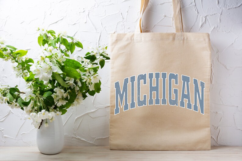 May include: A beige tote bag with the word "MICHIGAN" printed in blue on the front. The bag is sitting on a light wood surface in front of a white wall with a bouquet of white flowers in a white vase.