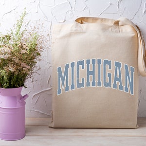 May include: A beige canvas tote bag with the word "MICHIGAN" printed in blue on the front. The bag is sitting on a wooden surface next to a pink metal vase with white flowers.