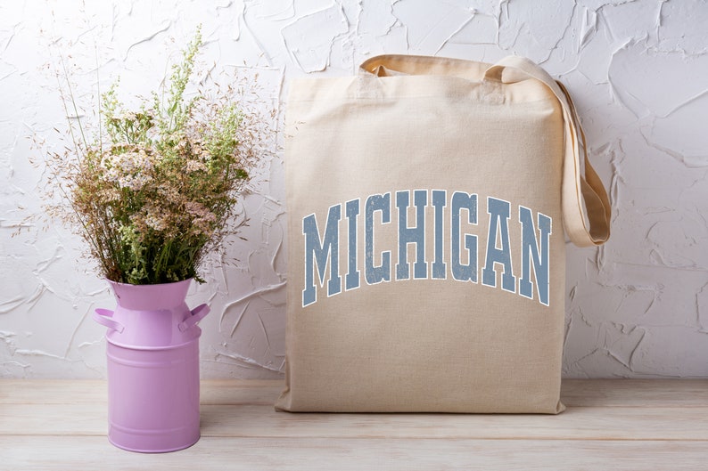 May include: A beige canvas tote bag with the word "MICHIGAN" printed in blue on the front. The bag is sitting on a wooden surface next to a pink metal vase with white flowers.