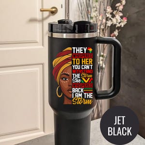 May include: A black travel mug with a handle and a straw. The mug features a graphic of a woman with a head wrap and the text "THEY WHISPERED TO HER YOU CAN'T WITHSTAND THE STORM She WHISPERED BACK I AM THE Storm". The mug is labeled "JET BLACK".