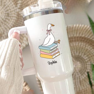 May include: White tumbler with a handle and a straw. The tumbler features a cartoon goose wearing a pink bow, sitting on a stack of colorful books. The name "Sophia" is printed below the illustration.