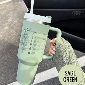 May include: Sage green tumbler with a handle and a white lid. The tumbler has the text "God says I am" and the name "SARAH" with associated words like "Strengthened" and "Abundant". A floral design is also present.