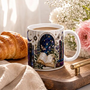 A white ceramic mug with a colorful, illustrated design sits on a wooden tray, accompanied by a croissant, a vase of white flowers, and a book.