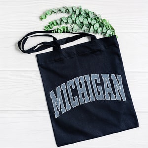 May include: A black tote bag with a white background and the word "MICHIGAN" printed in a distressed, gray font.