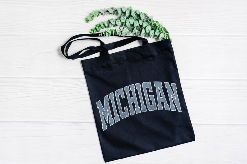 May include: A black tote bag with a white background and the word "MICHIGAN" printed in a distressed, gray font.
