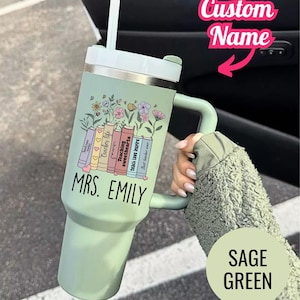 May include: Sage green tumbler with a handle and a white lid. The tumbler features a floral design with books and the text "Teacher life", "Teaching sweethearts", "teach love inspire", and "Best teacher ever". The name "MRS. EMILY" is also printed on the tumbler.