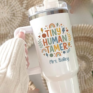 May include: White tumbler with a handle and a clear lid. The tumbler features the text "TINY HUMAN TAMER" in colorful letters, along with rainbows, lightning bolts, and a smiley face. The name "Mrs. Bailey" is printed below the text.