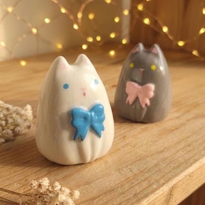 Può includere: Due statuette di gatto in ceramica. Una è bianca con un fiocco blu e occhi blu, l'altra è grigia con un fiocco rosa e occhi gialli. Le statuette di gatto sono su una superficie di legno.