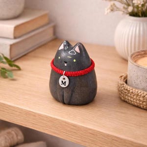 Op de afbeelding: Een glad, donkergrijs, afgerond stenen beeldje in de vorm van een kat. Het heeft roze geschilderde binnenoren, kleine gele ogen en een piepklein wit mondje. Een felrode gevlochten halsband omcirkelt de nek met een zilveren label met de letter M.
