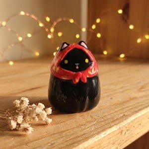 Può includere: Statuetta di gatto in ceramica nera con cappuccio rosso e dettagli dorati. Il gatto ha occhi gialli e una piccola lingua rosa. La statuetta è su una superficie di legno con fiori secchi.