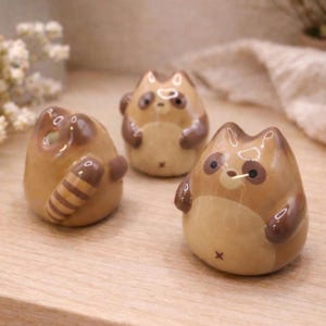 May include: Three ceramic raccoon figurines in tan and brown. Each figurine has a round body, small arms, and a raccoon tail. The raccoons have dark brown eyes and ears, and a light tan face and belly. The figurines are on a light wood surface.