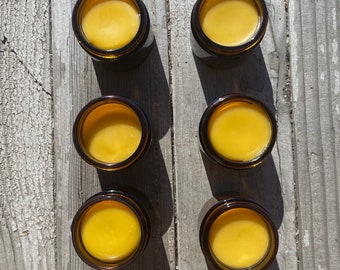 May include: Six amber glass jars filled with a yellow balm or cream. The jars are arranged in two rows of three on a light wood surface.