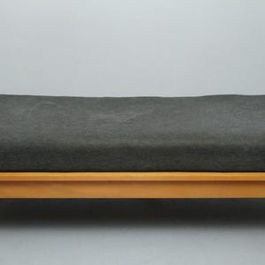 Può includere: Un divano letto rettangolare con un cuscino grigio scuro e una struttura in legno chiaro. Le gambe sono in metallo color argento. Il letto ha un design semplice e moderno.