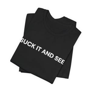 Puede incluir: Camiseta negra con el texto blanco "SUCK IT AND SEE" impreso en la parte delantera. La camiseta está doblada y parece estar hecha de un material suave. Se ve el cuello.