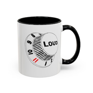 May include: A white ceramic mug with a black interior and handle. The exterior features a black line drawing of a circular volume control knob labeled with numbers one through eleven, with the word LOUD printed in black and the number eleven in red.