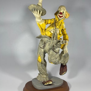 May include: A figurine of a clown with a yellow shirt, gray pants, and red shoes. The clown is holding a tray with a bottle on it. The figurine is on a wooden base.