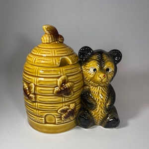 May include: A ceramic honey pot shaped like a beehive with a bear figurine next to it. The beehive has a brown glaze and is decorated with bees and leaves. The bear is brown and black with a red nose.