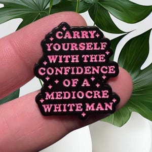 May include: A black and pink enamel pin with the text "CARRY YOURSELF WITH THE CONFIDENCE OF A MEDIOCRE WHITE MAN". The pin is held between fingers, with green leaves and white vases in the background.