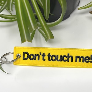 Puede incluir: Llavero rectangular amarillo con el texto bordado en negro "Don't touch me!". El llavero tiene un anillo y un cierre de metal plateado. Se ven hojas verdes de plantas en el fondo.