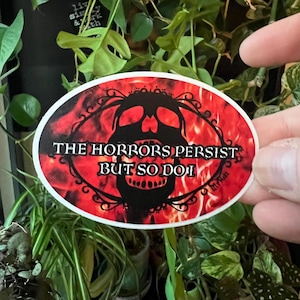 May include: Oval sticker with a red and black design. The sticker features a skull with the text "THE HORRORS PERSIST BUT SO DO I". The background is a fiery red, and the sticker is surrounded by green plants.