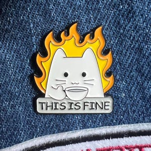 May include: A white cat with yellow flames on its head is holding a cup of coffee. The cat is sitting on a table with the text "This is fine" written below it.