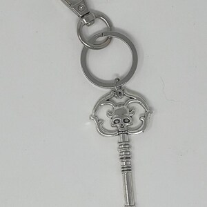 May include: Silver-colored key chain with a decorative key pendant. The key features a skull design at the top and a detailed handle. The key chain includes a split ring and a lobster clasp for attachment.