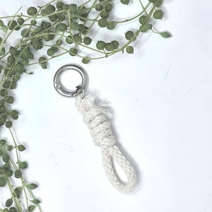May include: A close-up of a white rope keychain with a silver metal ring. The keychain is knotted at the top and has a loop at the bottom. Green plant stems with small round leaves are in the background.