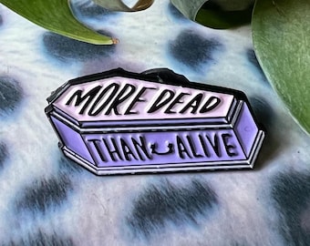 May include: A black and purple enamel pin shaped like a coffin with the text "More Dead Than Alive".