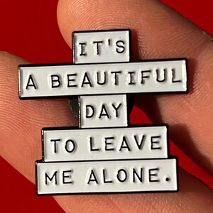 this is a funny introvert soft enamel pin that says "it's a beautiful day to leave me alone." This is a white pin with black lettering.