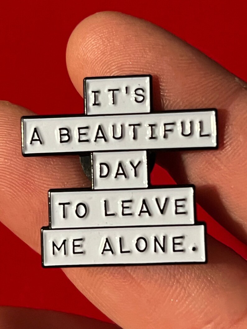 this is a funny introvert soft enamel pin that says "it's a beautiful day to leave me alone." This is a white pin with black lettering.