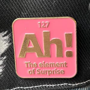 May include: A pink enamel pin with a gold border and gold lettering. The pin says "Ah! The element of Surprise" and has the number 127 above the word "Ah!"