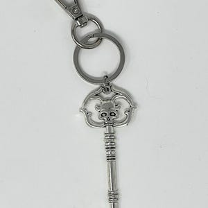 May include: Silver-colored key chain featuring a decorative key pendant with a skull design. The key has a detailed, ornate head and a long, textured stem. The key chain includes a split ring and a lobster clasp.