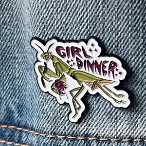 Pu&ograve; includere: Uno spillo in smalto bianco con una mantide religiosa verde che tiene un granchio rosso e viola. Lo spillo dice "Girl Dinner" in lettere rosa con stelle bianche.