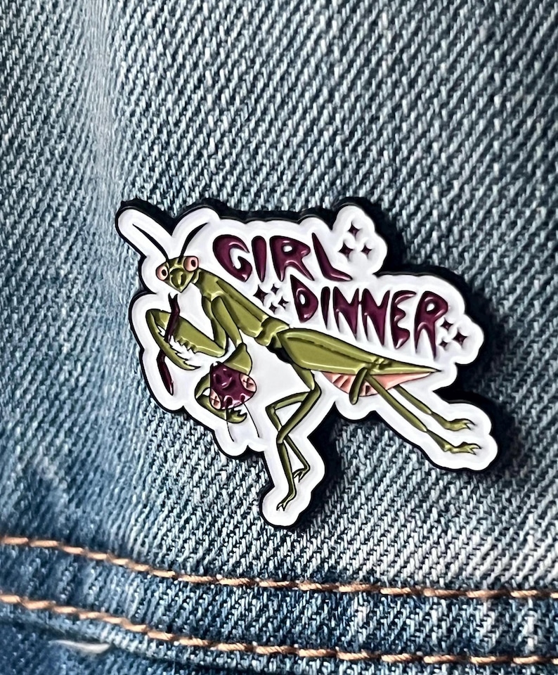 Pu&ograve; includere: Uno spillo in smalto bianco con una mantide religiosa verde che tiene un granchio rosso e viola. Lo spillo dice "Girl Dinner" in lettere rosa con stelle bianche.