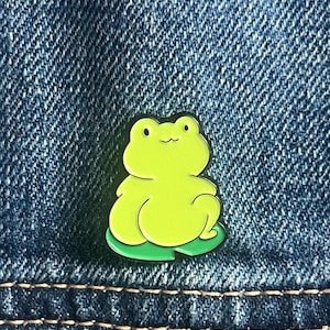 Puede incluir: Un pin de esmalte de rana verde lima con contorno negro, sentado sobre una hoja de nenúfar verde. El pin está colocado sobre un fondo de mezclilla azul.