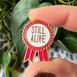 Peut inclure: Une épingle en émail de couleur dorée en forme de ruban rouge avec les mots "Still Alive" en lettres blanches sur un cercle blanc au centre.