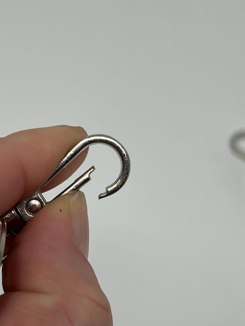 May include: Close-up of a silver-colored carabiner clip. The clip is open, revealing the spring-loaded mechanism. The background is a plain white surface.