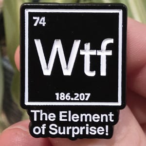 Op de afbeelding: Een zwart-witte rechthoekige pin met de tekst "Wtf" in grote witte letters. De pin bevat ook het nummer "74", "186.207" en de zin "The Element of Surprise!"