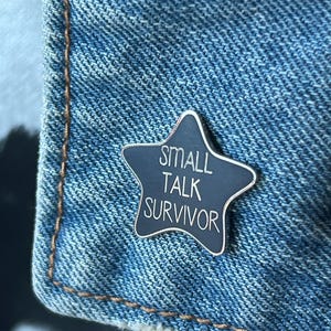 Puede incluir: Un pin de esmalte negro en forma de estrella con el texto "SMALL TALK SURVIVOR" en letras blancas.