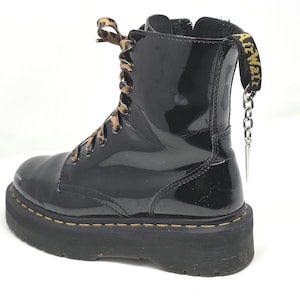 May include: Black platform boot with a glossy finish. The boot features leopard print laces and a yellow and black pull tab. A silver chain with a spike charm hangs from the pull tab. The sole has yellow stitching.
